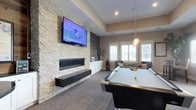 This single-floor space is designed as a sophisticated entertainment and recreation room, blending modern design elements with comfortable amenities. The focal point of the room is a striking stacked stone accent wall, which houses a sleek linear fireplace and a large wall-mounted flat-screen television, creating a dramatic and functional centerpiece. A floating dark shelf beneath the fireplace adds a contemporary touch to the natural stone texture.

The room features elevated ceilings with a tray ceiling detail, enhancing the sense of openness and volume. Recessed lighting is complemented by a stylish industrial-style pendant light that hangs above a slate-colored pool table, making it both a functional and decorative element.

Natural light enters through plantation-shuttered windows, contributing to the bright and airy feel of the space. A seating area near the windows, furnished with upholstered chairs accented with decorative pillows, provides a comfortable lounge zone. Built-in white cabinetry beneath the windows offers additional storage and display space. Neutral-toned carpet throughout ties the room together, creating a warm and inviting atmosphere suited for entertaining.