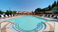 This outdoor amenity space centers around a generously sized resort-style swimming pool with crystal-clear blue water, accessible via wide entry steps equipped with safety handrails. The surrounding pool deck is finished in a light-toned, textured concrete with decorative circular inlay patterns, providing ample room for lounging. Rows of chaise lounge chairs and seating areas with tables and colorful market umbrellas in orange and yellow are arranged throughout the deck, creating a vibrant and welcoming atmosphere. A gracefully curved pergola structure with substantial square columns and an open-lattice roof runs along the perimeter of the space, casting dappled shade across the walkway beneath. The pergola connects to an ornamental black wrought iron fence and gate, which encloses the area while maintaining open sightlines to surrounding greenery. Carefully maintained landscaping features ornamental grasses, flowering shrubs, and young trees set against a stone retaining wall, adding natural texture and color throughout. The multi-story building visible in the background provides architectural context, while the open sky above ensures the space is bathed in natural light throughout the day, creating an inviting outdoor retreat.