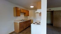 This single-floor residential space offers a practical and straightforward layout designed with everyday functionality in mind. The kitchen is a central feature, showcasing warm-toned wood cabinetry that provides generous storage through both upper and lower cabinets and drawers. Light countertops offer ample workspace and are complemented by a stainless single-basin sink. White appliances, including a coil-burner electric range with an overhead ventilation hood and a full-sized refrigerator, complete the kitchen setup. A textured ceiling adds a subtle visual layer to the space, while flush-mounted ceiling lighting ensures the kitchen remains well-lit. The kitchen opens toward an adjoining area, suggesting a connected and accessible flow between living spaces. The flooring transitions from durable, dark tile in the kitchen to carpeted areas beyond, offering a practical distinction between functional and relaxation zones. The overall ambiance is clean, utilitarian, and move-in ready, making it a solid and comfortable space suited for practical, everyday living.