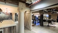 This single-floor space masterfully blends industrial and rustic design elements to create a distinctive, characterful environment. Structural brick columns — some in classic red brick, others in natural stacked stone — serve as architectural focal points throughout, adding texture and a sense of history to the open layout. Exposed ceiling beams and visible ductwork reinforce the industrial aesthetic, while recessed and track lighting ensures the space is well-lit and functional.

A standout feature is the sliding barn door, crafted from weathered wood with cross-brace detailing and mounted on black hardware, which opens into a dedicated recreation area. This lounge space is anchored by a dark-wood billiards table positioned beneath a dramatic cluster chandelier of globe pendants, creating an inviting and social atmosphere. Light tile flooring runs throughout, providing a neutral contrast to the warm tones of the brick, wood, and stone. White cylindrical columns blend seamlessly with the modern finishes, balancing the rustic elements with a cleaner, contemporary touch. The overall ambiance is one of thoughtfully layered design — combining raw materials and refined finishes to deliver a space that is both visually engaging and highly functional.