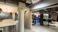 This single-floor space masterfully blends industrial and rustic design elements to create a distinctive, characterful environment. Structural brick columns — some in classic red brick, others in natural stacked stone — serve as architectural focal points throughout, adding texture and a sense of history to the open layout. Exposed ceiling beams and visible ductwork reinforce the industrial aesthetic, while recessed and track lighting ensures the space is well-lit and functional.

A standout feature is the sliding barn door, crafted from weathered wood with cross-brace detailing and mounted on black hardware, which opens into a dedicated recreation area. This lounge space is anchored by a dark-wood billiards table positioned beneath a dramatic cluster chandelier of globe pendants, creating an inviting and social atmosphere. Light tile flooring runs throughout, providing a neutral contrast to the warm tones of the brick, wood, and stone. White cylindrical columns blend seamlessly with the modern finishes, balancing the rustic elements with a cleaner, contemporary touch. The overall ambiance is one of thoughtfully layered design — combining raw materials and refined finishes to deliver a space that is both visually engaging and highly functional.
