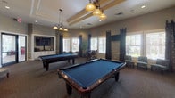 This single-floor recreational space is thoughtfully designed to serve as a social hub for entertainment and leisure. The room makes an immediate impression with its bold, decorative damask-patterned wallpaper in warm gold and soft blue tones, lending the space a refined yet lively character.

At the heart of the room, two full-size billiard tables with rich hardwood frames and blue felt surfaces take center stage, each illuminated by elegant pendant light fixtures with wrought iron detailing. The tray ceilings add architectural depth and a sense of elevated formality to the space.

An arcade area along one wall features several upright gaming cabinets, including a pinball machine and video game stations, catering to a variety of recreational interests. Comfortable upholstered accent chairs with soft patterned fabric are arranged nearby, providing inviting spots to relax between activities.

A high-top table with bar-height chairs is positioned near the windows, which allow natural light to filter in through slatted blinds framed by dramatic dark curtain panels. Tall decorative indoor plants add organic warmth throughout.

A wall-mounted flat-screen television and additional seating areas complete the lounge atmosphere, while large windows offer views of an adjacent outdoor area. The neutral carpet, recessed lighting, and cohesive décor tie the entire space together into a welcoming, well-appointed recreational venue.