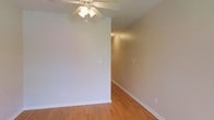 This two-floor space presents a straightforward, functional layout with a clean and neutral aesthetic throughout. The main living area, visible in the provided image, features warm-toned hardwood flooring that adds a natural element to the space and complements the soft, beige-toned walls. A ceiling fan with an integrated light fixture is centrally positioned overhead, providing both air circulation and ambient lighting. White baseboards run along the perimeter of the room, offering a crisp contrast against the flooring and walls. The open sightlines visible through the hallway suggest a connected floor plan that allows for natural flow between areas. The walls are smooth and move-in ready, providing a blank canvas for personalization. The overall ambiance of the space is calm and neutral, suited to a variety of furnishing styles. With two floors, the property offers opportunities for distinct living and private zones, though specific details of the upper level are not shown. The space presents a clean, well-maintained environment with functional design elements throughout.