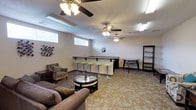 The Resident Lounge is a generously sized, single-floor communal space designed to provide residents with a comfortable and versatile gathering area. The lounge is laid out across an open floor plan with patterned carpeting throughout, lending a warm and cohesive aesthetic to the entire space.

A central highlight is the built-in bar counter, complete with a dark countertop and equipped with bright green adjustable barstools, making it an ideal spot for casual socializing. Adjacent to the bar, additional seating options include a triangular accent table and a tall open bookshelf for display or storage.

Multiple lounge seating arrangements are distributed throughout the space, featuring plush sofas, cushioned armchairs, and coffee tables that invite relaxed conversation or quiet reading. Decorative wall art adds character to the neutral-toned walls, while a Coca-Cola vending machine provides convenient refreshments for residents.

Natural light filters in through horizontal slit windows positioned along the upper portion of the walls. Multiple ceiling fans with integrated lighting fixtures ensure consistent air circulation and illumination throughout. A separate, quieter nook offers additional seating with a classic wooden coffee table and accent chair, ideal for reading or casual meetings.