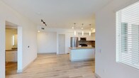 Located at West Ranch in Carlsbad, this single-floor unit offers a clean, modern aesthetic with an open-concept design that seamlessly connects the living and kitchen areas. Light hardwood flooring flows throughout the space, enhancing the bright and airy atmosphere created by recessed lighting and a track lighting fixture in the living area.

The kitchen is a standout feature, boasting white cabinetry, a tasteful tile backsplash, and a suite of stainless steel appliances including a refrigerator, microwave, and range. A central island with a neutral countertop provides additional workspace and casual seating, while elegant pendant lights hang above, adding a contemporary finishing touch. White upper cabinetry extends to the ceiling, maximizing storage while maintaining a sleek, cohesive look.

The open living area offers generous space for flexible furniture arrangements and is bathed in ambient light from the well-planned interior lighting. Crisp white walls throughout the unit contribute to a fresh, move-in-ready feel. The overall design balances modern functionality with a welcoming ambiance, making this Carlsbad residence a well-appointed and thoughtfully designed space.