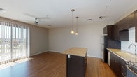 This single-floor residence features a contemporary open-concept layout that seamlessly connects the kitchen and living area. The kitchen is well-appointed with dark espresso cabinetry extending from floor to ceiling, granite countertops, a white subway tile backsplash, and a full suite of stainless steel appliances including a refrigerator, range, microwave, and dishwasher. A central island with matching granite provides additional workspace and casual seating, illuminated by a pair of pendant lights overhead. Track lighting and recessed vents complement the clean ceiling design throughout. The living area is generously sized with warm hardwood-style flooring, neutral gray walls, and a ceiling fan. A full-length sliding glass door with vertical blinds opens to an outdoor view and fills the space with natural light. The bedroom features plush carpeting, a ceiling fan with light fixture, and a window with blinds. The bathroom is finished consistently with the rest of the unit, showcasing dark cabinetry with a granite vanity, a tub-shower combination with tiled surround, and hardwood-style flooring, creating a cohesive and modern aesthetic throughout the space.