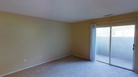 This single-floor space presents a straightforward and functional layout highlighted by clean, minimal design. The room visible in the images features wall-to-wall carpeting in a neutral grey-beige tone, providing a soft and comfortable underfoot feel throughout. The walls are finished in a warm yellow hue, contributing a subtle sense of warmth to the interior. White trim lines the base of the walls, offering a clean contrast against both the flooring and wall color.

A notable feature of this room is the sliding glass door positioned along one wall, framed by sheer white curtains that diffuse incoming natural light. The door provides direct access to an outdoor area, allowing for ventilation and outdoor views. The natural light filtering through the sheer panels brightens the interior, enhancing the room's open and airy feel.

The ceiling is finished in white and sits at a standard height, maintaining a clean and uncluttered overhead appearance. A ceiling vent is visible, indicating a climate control system is in place. The overall ambiance of the space is calm and neutral, offering a versatile backdrop suitable for a variety of uses and furnishing styles.