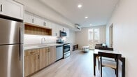 This thoughtfully designed single-floor unit offers a clean, modern aesthetic across four distinct spaces. The open-concept kitchen and living area serves as the heart of the unit, featuring a fully equipped kitchen with white upper cabinetry, warm wood-toned lower cabinets, stainless steel appliances including a refrigerator, electric range, built-in microwave, and dishwasher, as well as under-cabinet lighting and a single-basin sink. Light hardwood flooring flows seamlessly throughout, lending warmth and continuity to the space. The adjoining living area is bright and inviting, benefiting from large windows with distinctive decorative lattice panels that allow natural light to fill the room while offering urban views. A wall-mounted mini-split system provides efficient climate control. Two well-proportioned bedrooms each feature light wood platform bed frames and clean white walls, with windows continuing the natural light theme seen throughout. The bathroom is finished in crisp white, featuring a full soaking tub and shower surround with recessed lighting above. Recessed ceiling lights and a neutral color palette throughout create a cohesive, contemporary atmosphere that is both functional and visually refined.