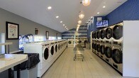 This single-floor laundromat offers a clean, efficiently organized space designed to accommodate a high volume of laundry needs. The main area is lined on both sides with rows of commercial-grade front-loading washing machines and stacked dryers, providing ample capacity for customers. The flooring consists of large, light-colored tiles that contribute to the bright, open feel of the space. Overhead, a combination of recessed lighting and a striking row of globe pendant lights runs the length of the room, ensuring thorough illumination throughout. A bold blue chevron-patterned accent wall at the far end serves as a distinctive design feature, adding visual interest and a modern aesthetic. Gray painted walls complement the overall contemporary style. Rolling laundry carts are available in the central aisle for customer convenience, and a mounted flat-screen television is positioned toward the rear of the space, providing entertainment during wait times. Natural light filters in through windows along one side of the facility. The layout maximizes functionality while maintaining a tidy, welcoming atmosphere, making it a practical and well-appointed commercial laundry facility.