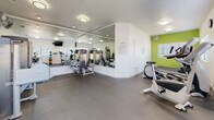 Located at Avenida Alcachofa in San Diego, CA 92128, this single-story fitness center offers a comprehensive workout environment designed to support a variety of exercise routines. The space is finished with durable dark flooring throughout, providing a practical and clean surface suited for both cardio and strength training activities.

The facility is divided into distinct workout zones. One area houses a selection of strength-training machines, positioned in front of expansive floor-to-ceiling mirrors that visually enlarge the space and allow users to monitor their form. A vibrant lime-green accent wall adds a bold, energizing contrast to the otherwise neutral white walls and ceiling. Ceiling-mounted flush lighting ensures consistent illumination across the entire facility.

The cardio zone is well-appointed with elliptical trainers, stationary bikes, and a treadmill, complemented by a wall-mounted flat-screen television for entertainment during workouts. Multiple windows fitted with horizontal blinds allow natural light to filter in while maintaining privacy. Additional functional fitness equipment, including kettlebells, medicine balls in various weights, and a stability ball, rounds out the offerings. A glass-panel entry door provides easy outdoor access, contributing to an open and welcoming atmosphere throughout the space.