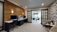 This single-floor business center combines contemporary design with functional workspaces, creating an environment well-suited for both focused individual work and group collaboration. The main workspace area is anchored by a built-in desk station equipped with multiple computers and a multifunction printer, set against a striking accent wall clad in warm, natural wood planking. Wall sconces provide ambient lighting, complementing the recessed ceiling lights throughout.

A standout feature is the pair of industrial-style black-framed sliding barn doors with frosted glass panels, which open to reveal a dedicated conference room. This private meeting space showcases a live-edge wood conference table surrounded by modern upholstered chairs, with floor-to-ceiling windows offering abundant natural light and views of the surrounding exterior. The lounge area features plush, textured seating arranged alongside a decorative tile accent wall finished in layered metallic tones, adding visual depth and sophistication.

Additional details include a private office or secondary workstation room, decorated with curated artwork and accessible via a dark-framed entry door. Soft, neutral carpeting runs throughout, tying the various zones together cohesively. The overall design aesthetic blends warm organic materials with sleek modern finishes, resulting in a polished and professional atmosphere.