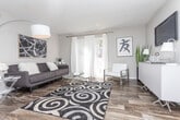 This single-floor residence offers a thoughtfully designed layout that balances functionality with contemporary style. The living room serves as a welcoming central space, featuring wood-look flooring, neutral gray walls, and a large window dressed with sheer white curtains that allow natural light to fill the room. A bold abstract artwork anchors the wall above the seating area, complementing the modern aesthetic.

The kitchen is well-equipped with stainless steel appliances, including a refrigerator, range, and dishwasher, set against white cabinetry with brushed nickel hardware. A wood-toned tile backsplash adds texture, while light countertops provide ample workspace. The kitchen opens toward a dedicated dining area, which features wood-look flooring and room for a small round table.

Two bedrooms offer carpeted flooring, natural light through blinded windows, and neutral wall tones. The primary bedroom is generously sized and includes access to a walk-in closet, while both rooms provide a calm, restful ambiance. Throughout the unit, a cohesive palette of grays, whites, and warm wood tones creates a clean, modern atmosphere.