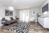 This single-floor residence offers a thoughtfully designed layout that balances functionality with contemporary style. The living room serves as a welcoming central space, featuring wood-look flooring, neutral gray walls, and a large window dressed with sheer white curtains that allow natural light to fill the room. A bold abstract artwork anchors the wall above the seating area, complementing the modern aesthetic.

The kitchen is well-equipped with stainless steel appliances, including a refrigerator, range, and dishwasher, set against white cabinetry with brushed nickel hardware. A wood-toned tile backsplash adds texture, while light countertops provide ample workspace. The kitchen opens toward a dedicated dining area, which features wood-look flooring and room for a small round table.

Two bedrooms offer carpeted flooring, natural light through blinded windows, and neutral wall tones. The primary bedroom is generously sized and includes access to a walk-in closet, while both rooms provide a calm, restful ambiance. Throughout the unit, a cohesive palette of grays, whites, and warm wood tones creates a clean, modern atmosphere.