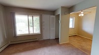 This single-floor, one-bedroom, one-bathroom residence offers a well-organized layout designed for comfortable everyday living. The living area features neutral-toned carpeting, taupe walls, and a large sliding window that brings in natural light while offering outdoor views of surrounding greenery. A louvered closet door and entry door are conveniently positioned in this space, with a baseboard heating unit running along the exterior wall. An adjoining area, also carpeted and bathed in natural light from a second window, provides additional flexibility for use as a dining or sitting space.

The bedroom is a generously sized, carpeted room with bright white walls, a sliding window, and its own baseboard heater. A wall-mounted air conditioning unit adds year-round climate control. A built-in closet with louvered double doors provides ample storage with a full-width hanging rod and shelf.

The galley-style kitchen is a standout feature, equipped with upgraded oak cabinetry lining both sides, a light-toned countertop, and a suite of black appliances including a refrigerator, over-the-range microwave, electric range, and dishwasher. Tile flooring completes the space.

The bathroom includes a tub-shower combination with white tile surround, a window for natural ventilation, tile flooring, and an oak vanity with a marble-style countertop.