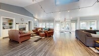 This single-story space in Boise, Idaho, is designed as a welcoming communal environment with a thoughtful, open layout. The main gathering area is defined by soaring vaulted ceilings with track lighting that illuminates the expansive hardwood floors throughout. Soft blue-gray walls and crisp white trim create a clean, inviting aesthetic. A built-in shelving unit flanks a wall-mounted television and fireplace, providing a comfortable focal point for the seating area, which features leather sofas and armchairs arranged on area rugs. Large windows and glass-paneled French doors along one wall allow abundant natural light to pour in, offering views of the surrounding greenery and an adjacent deck. A casual dining nook positioned near the windows accommodates a small round table with vibrant teal chairs, adding a pop of color to the space. The kitchen features sleek white cabinetry, granite-style countertops with a mosaic tile backsplash, and ample counter space, with multiple windows framing outdoor views. A dedicated computer room off the main area provides a quieter workspace with built-in desks, computers, and storage cabinetry, finished with carpeted flooring. Overall, the space balances functionality with a relaxed, modern character.