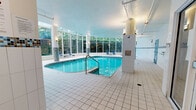 Located at 3551 Boulevard Gouin Est in Montréal, Québec, this indoor aquatic facility offers a well-appointed and generously sized pool area spread across a single level. The centerpiece of the space is a large, free-form indoor swimming pool with vivid turquoise water, complemented by a separate whirlpool/hot tub positioned nearby. The pool surround is finished in clean, white large-format tiles, providing a bright and hygienic deck area with ample space for movement around the water.

A defining architectural feature is the expansive floor-to-ceiling glazed wall that spans one full side of the pool area, allowing an abundance of natural light to flood the interior while framing lush, tree-lined outdoor views. This greenhouse-style glazing creates an open, airy ambiance that balances the enclosed nature of an indoor facility.

The interior walls are finished with white tile accented by a decorative mosaic border in multicoloured tones, adding a visual detail consistent throughout the space. Structural columns adorned with matching mosaic tile provide both support and design continuity. Bold blue accent doors punctuate the white walls, adding a striking contrast. Access points including a glass-enclosed entry vestibule and stainless steel pool entry rails are also present, ensuring accessibility. The facility is well-lit with wall-mounted fixtures complementing the natural daylight throughout.