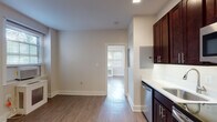 This single-floor unit offers a clean, modern layout thoughtfully designed for everyday living. The kitchen is a standout feature, showcasing rich dark espresso cabinetry that extends floor to ceiling, white quartz countertops, a stainless steel undermount sink, and a full suite of stainless steel appliances including a dishwasher, electric range, and over-the-range microwave. Recessed lighting overhead adds a polished finishing touch to the cooking space.

The open living area flows seamlessly from the kitchen, featuring warm hardwood-style plank flooring that runs throughout the unit. Neutral beige walls create a bright and versatile backdrop, while large windows fitted with horizontal blinds allow abundant natural light to fill the space.

The bedroom is generously proportioned, offering a wide bank of windows with outdoor views and a set of double doors leading to a closet. A radiator unit beneath the windows provides efficient in-room heating.

The bathroom is elegantly detailed with classic black-and-white octagonal tile flooring, white subway tile wainscoting, a marble-topped vanity with dark wood cabinetry, and a small window that introduces natural light. The overall ambiance throughout the unit is clean, bright, and move-in ready, with a cohesive design palette that balances modern finishes with timeless details.