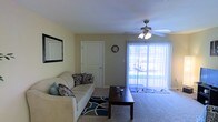 This single-floor residence features a welcoming living room that serves as the central gathering space. The room is finished with wall-to-wall carpeting and painted in a warm neutral tone, contributing to an inviting and relaxed ambiance. A ceiling fan with an integrated light fixture is centrally mounted, providing both airflow and general illumination. Natural light flows generously into the space through a large sliding glass door dressed with vertical blinds, offering a view of the outdoors and brightening the interior throughout the day. The living area is furnished with a cushioned sofa accented by decorative throw pillows, paired with a dark wood coffee table set atop a patterned area rug. A wall-mounted mirror adds visual depth to the space. An interior door with a classic paneled design leads to an adjacent room, suggesting additional living areas beyond. A decorative wall clock adds a functional accent to the neutral walls. Overall, the space presents a practical and comfortable layout with a clean, straightforward design well-suited for everyday living.