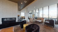 This sophisticated single-floor amenity space is thoughtfully designed to accommodate both relaxed socializing and casual dining. The lounge area anchors one end of the space, featuring plush sectional sofas, leather barrel chairs, and sleek accent seating arranged around a modern coffee table. A dramatic floor-to-ceiling stone fireplace serves as a striking focal point, while expansive curtain-wall windows flood the room with natural light and frame sweeping panoramic city skyline views.

The dining and kitchen zone is equally well-appointed, showcasing a bold mosaic tile feature wall behind a built-in bar and television. Pendant lighting in matte black and metallic finishes hang over a white quartz bar counter lined with wood-and-metal counter stools. Patterned tile flooring defines this area, complemented by dark hardwood-style flooring throughout the dining space, which offers multiple seating configurations including communal tables and intimate banquette booths.

A connecting corridor provides a transitional element between zones, featuring reclaimed wood slat partitions, dark hardwood flooring, and curated artwork along charcoal-toned walls. The overall design palette blends warm wood tones, rich textiles, metallic accents, and neutral upholstery to create an inviting, upscale atmosphere suited for both quiet relaxation and lively gatherings.