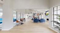 This single-floor community clubhouse is thoughtfully designed to serve as a welcoming gathering space, combining open-plan living with modern, curated interiors. The main lounge area commands attention with soaring vaulted ceilings, a striking geometric chandelier, and a floor-to-ceiling white brick fireplace that anchors the room. Blue velvet sofas and armchairs are arranged around a live-edge wood coffee table atop a striped area rug, creating an inviting atmosphere for socializing or relaxing. Light-toned hardwood flooring runs throughout, amplifying the sense of openness and warmth.

A gallery wall of bold, colorful abstract artwork adds a vibrant focal point, while tall windows and glass French doors flood the space with natural light and offer views of an outdoor pool area. A casual dining zone with round tables and leather chairs provides a more intimate seating option.

The kitchen features white shaker cabinetry, light quartz countertops, a subway tile backsplash, and stainless steel appliances including a refrigerator, microwave, and sink, making it well-suited for events and gatherings. A reception and workstation area with computer desks and bar-height seating rounds out the space, lending it a functional, multi-purpose character throughout.