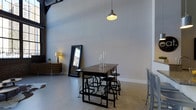 This single-floor space showcases a refined industrial aesthetic, combining raw architectural elements with thoughtfully curated furnishings. The most striking feature is the impressively high ceiling, which lends the interior a dramatic sense of openness and volume. Polished concrete floors run throughout, providing a clean, modern foundation that complements the industrial character of the space.

The main area is anchored by a dark-toned industrial dining or work table paired with adjustable metal stools, reflecting a functional yet stylish sensibility. A single pendant light with a utilitarian shade hangs overhead, casting warm, focused light over the central gathering area. Along one wall, a wooden sideboard adds a touch of warmth and storage, accompanied by a floor lamp that softens the ambiance. A large, black-framed floor mirror leans against the wall, visually expanding the space while hinting at natural light from nearby windows.

The neutral grey walls and pale concrete floors create a cohesive, understated palette that allows the architectural scale and carefully chosen furnishings to take center stage. Exposed structural elements further reinforce the industrial character. Overall, the space conveys a cool, contemporary atmosphere well-suited for both productive work and relaxed social gatherings.