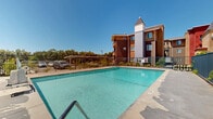 Located at 975 14th St in Oroville, California, this outdoor pool area is a standout communal amenity set against a backdrop of multi-story residential buildings. The centerpiece is a large rectangular swimming pool with clear, blue-tinted water, designed to accommodate both leisure and lap swimming. The pool is framed by a wide, paved pool deck that provides ample space for lounging and socializing under the California sun. A secure wrought-iron perimeter fence encloses the entire area, ensuring safety and privacy for users. Entry steps and handrails offer convenient access into the pool from multiple points. Surrounding the deck, carefully maintained shrubs and greenery add a touch of natural beauty to the space. The adjacent multi-story buildings feature warm earth-toned exteriors with distinctive peaked rooflines, cream-colored column accents, and private balconies that overlook the pool area. Open sky views complete the setting, making this an inviting outdoor retreat. The 3D scan reveals the full footprint of the pool enclosure, highlighting the generous scale of the deck and the well-defined boundaries of this amenity space.