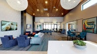 This contemporary amenity lounge is thoughtfully designed to blend comfort, style, and functionality into a single, open-concept space. The standout architectural feature is the rich, warm-toned wood-paneled ceiling, which adds texture and elegance while contrasting beautifully with the clean white walls and modern finishes throughout. Statement globe pendant lights hang at varying heights, casting a soft, inviting glow across the room.

The lounge area is furnished with plush navy blue sofas and accent chairs adorned with colorful throw pillows, creating a relaxed yet sophisticated seating arrangement centered on a patterned area rug. Adjacent to the lounge, a full-size billiards table serves as a focal point for recreation and social interaction.

Bar-height seating with teal upholstered chairs lines a counter area, offering a more casual dining or social setting. A sliding barn door adds a rustic-modern touch, partially visible near what appears to be a secondary room or workspace. Large floor-to-ceiling windows and glass doors flood the space with natural light and provide open sightlines to surrounding areas, enhancing the sense of airiness. The durable wood-look flooring ties all zones together seamlessly, making this lounge a versatile and welcoming communal destination.