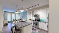 This single-story residence offers a thoughtfully designed open-concept layout where the kitchen flows seamlessly into the living area. The kitchen serves as a central highlight, showcasing a large island with striking dark granite countertops, an undermount sink, and seating for casual dining. White shaker-style cabinetry with glass-front uppers, a soft green subway tile backsplash, and stainless steel appliances—including a range, over-the-range microwave, and refrigerator—complete the space. Track lighting and globe pendant fixtures provide layered illumination throughout.

The living area features warm hardwood-style flooring, a soothing teal accent wall, and floor-to-ceiling French doors that invite generous natural light. A ceiling fan adds comfort to this welcoming gathering space.

The bedroom is carpeted and comfortably scaled, with dual windows offering natural light, a ceiling fan, and direct access to the en-suite bathroom. The bathroom features a double-sink vanity with dark espresso cabinetry, granite countertops, a large framed mirror, and a separate water closet with a walk-in shower and tub combination. The overall palette is modern and cohesive, with neutral tones accented by teal and warm wood finishes throughout.