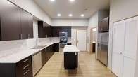 This single-floor, one-bedroom, one-bathroom residence offers a well-planned layout designed for comfortable, modern living. The kitchen serves as a central highlight, featuring dark espresso cabinetry that extends to the ceiling, contrasted beautifully by light marble-style countertops. A central island provides additional prep space and casual seating, making it ideal for both cooking and entertaining. Stainless steel appliances — including a French door refrigerator, electric range with overhead microwave, and dishwasher — complete the kitchen's contemporary and functional aesthetic. Light wood-tone luxury vinyl flooring runs throughout, lending warmth and visual continuity across the space. Recessed lighting illuminates the kitchen evenly, creating a bright and clean atmosphere. A doorway visible from the kitchen leads toward the bathroom, which features dark cabinetry consistent with the kitchen's design language. The overall ambiance is sleek and modern, with a neutral color palette of whites and grays complemented by the rich, dark tones of the cabinetry. The open flow between spaces maximizes the sense of room and ensures ease of movement throughout the residence.