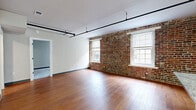 Located at 12 South 17th Street in Richmond, VA, this one-bedroom, one-bathroom unit masterfully combines historic architectural charm with contemporary updates. The open-concept kitchen and living area serves as the heart of the space, where warm hardwood floors stretch beneath striking exposed brick walls punctuated by large windows that flood the room with natural light. The kitchen is well-appointed with white shaker cabinetry, granite countertops, stainless steel appliances, and a central island with a built-in sink — all framed by dramatic rustic wood beams overhead that speak to the building's storied past. The living area offers an airy, loft-like ambiance enhanced by sleek black track lighting along the ceiling. The bedroom features plush carpeting and generous double closets with bifold doors, providing ample storage. The full bathroom is finished with large-format tile flooring, a white vanity with granite countertops, a framed mirror, and a tub-shower combination. A dedicated laundry area and entrance hallway complete the layout. This thoughtfully designed space balances the raw character of exposed brick with polished modern finishes throughout.