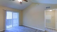 This single-floor space is defined by its vaulted ceiling, which lends a sense of openness and volume to the main living area. The angled ceiling rises to a notable peak, enhancing the airy quality of the room and creating a welcoming sense of spaciousness. A ceiling fan with a wood-toned finish is centrally mounted, providing both functional air circulation and a subtle decorative element.

The flooring throughout is covered in plush, neutral-toned carpeting, offering a soft and comfortable surface underfoot. The walls are finished in a warm, light yellow tone that complements the natural light filtering into the space. Vertical blinds cover a sliding glass door along one wall, which serves as the primary source of natural light and likely provides access to an outdoor area. When open, the blinds allow generous daylight to brighten the interior.

White baseboards frame the perimeter of the room, providing a clean contrast against the carpeted floor and painted walls. Electrical outlets are positioned at standard intervals along the walls. Overall, the space presents a clean, functional layout with architectural details that contribute to a bright and comfortable atmosphere.