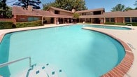 This outdoor pool area offers a generously sized swimming pool with clear, turquoise water and tiled accents along the pool floor, providing a refreshing and visually inviting centerpiece. The pool is bordered by a wide, smooth concrete deck that offers ample space for lounging and movement around the water. Safety features include clearly marked depth indicators along the pool edge and accessible entry points with metal handrails.

The surrounding area is enclosed by a classic combination of brick columns and black iron fencing, creating a secure and well-defined boundary. Brick-pillared gated entry points provide controlled access to the pool area. Outdoor seating is available on the deck, with table-and-chair sets positioned under shade umbrellas, offering a comfortable spot to relax.

Mature trees lining the perimeter provide natural shade and greenery, enhancing the outdoor ambiance. Landscaped beds with shrubs and ground cover add to the well-maintained appearance of the space. Trash receptacles are conveniently located within the enclosure. The backdrop features multi-story residential buildings with brick exteriors and pitched rooflines, integrating the pool area seamlessly into the broader community. Under open skies, this pool area serves as a functional and welcoming outdoor retreat.