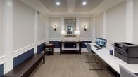 This single-floor business center is thoughtfully designed with a blend of traditional elegance and functional workspace elements. The room's architectural character is defined by white wainscoting and panel molding that frames the walls, with a rich navy blue lower half creating a striking two-tone contrast against the soft cream upper walls. Recessed lighting and a tray ceiling add refinement overhead, while neutral-toned carpet grounds the space comfortably underfoot.

Along one wall, a built-in workstation features a clean white countertop surface equipped with computers and a printer, providing a practical workspace for multiple users simultaneously. Complementing this is a dark wood console table centered against the far wall, flanked by classic wall sconces and topped with matching table lamps, with a gold-framed mirror above lending a decorative focal point. A dark leather bench and a cherry wood accent table with a glass inset offer additional seating and surface space.

Large multi-pane windows along one side allow natural light to filter in, offering views into adjacent common areas. An open doorway connects seamlessly to neighboring spaces, enhancing the overall sense of accessibility and flow throughout the building.
