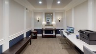 This single-floor business center is thoughtfully designed with a blend of traditional elegance and functional workspace elements. The room's architectural character is defined by white wainscoting and panel molding that frames the walls, with a rich navy blue lower half creating a striking two-tone contrast against the soft cream upper walls. Recessed lighting and a tray ceiling add refinement overhead, while neutral-toned carpet grounds the space comfortably underfoot.

Along one wall, a built-in workstation features a clean white countertop surface equipped with computers and a printer, providing a practical workspace for multiple users simultaneously. Complementing this is a dark wood console table centered against the far wall, flanked by classic wall sconces and topped with matching table lamps, with a gold-framed mirror above lending a decorative focal point. A dark leather bench and a cherry wood accent table with a glass inset offer additional seating and surface space.

Large multi-pane windows along one side allow natural light to filter in, offering views into adjacent common areas. An open doorway connects seamlessly to neighboring spaces, enhancing the overall sense of accessibility and flow throughout the building.