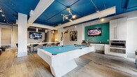 Located at the NC Music Factory in Charlotte, NC, this single-floor entertainment lounge delivers a dynamic, music-inspired atmosphere with a distinct urban edge. The space is anchored by a bold cobalt-blue ceiling that contrasts against white structural beams and exposed ductwork, lending an industrial-chic aesthetic throughout. Wide-plank wood-look flooring runs seamlessly across the entire floor, grounding the energetic design.

At the heart of the space sits a sleek white pool table with blue felt, positioned beneath a sculptural pendant light fixture that serves as a striking focal point. Multiple flat-screen TVs are mounted throughout, including one set against a teal mosaic tile accent wall, ensuring entertainment from every angle. A high-top bar counter with blue upholstered stools provides additional gathering space, while a lounge area with a brown leather sofa and accent chairs offers a more relaxed setting.

Bold large-scale murals on glass-paneled doors and walls add an artistic, gallery-like quality to the space. Music-themed décor — including a wall-mounted guitar, framed portraits, and eclectic wall art — reinforces the cultural identity of the NC Music Factory setting. Track lighting and pendant fixtures illuminate the space with a warm, inviting glow, making this lounge a well-rounded venue for socializing and entertainment.