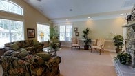 Located at 901 Falls Park Dr in Sanford, NC, this single-floor space offers a refined and welcoming atmosphere with carefully considered interior design. The main sitting area is generously proportioned, featuring soft carpeted flooring and warm, neutral-toned walls that create a calm and comfortable ambiance. Recessed lighting in the ceiling provides even, pleasant illumination throughout the space, complemented by natural light streaming in through large windows dressed with white plantation shutters. The windows offer views of surrounding greenery, bringing the outdoors in and enhancing the bright, airy feel of the room. Elegant crown molding adds a classic architectural detail that frames the ceiling with sophistication. The furnishings include richly upholstered floral-patterned seating alongside traditional striped wingback chairs, lending the space a timeless, classic character. Decorative touches such as tall indoor tropical plants, framed wall art, and a glass-top accent table with tasteful centerpieces contribute to the polished, curated aesthetic. The overall ambiance is one of quiet elegance and comfort, making this a welcoming and well-appointed space suited for relaxed gathering and refined living.