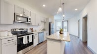 This single-floor residence showcases a thoughtfully designed open layout with consistent wood-style flooring flowing throughout the main living areas. The kitchen serves as a central highlight, featuring crisp shaker-style cabinetry in a soft grey-white finish, granite countertops, a white subway tile backsplash, and a full suite of stainless steel appliances including a range, microwave, dishwasher, and refrigerator. A spacious kitchen island with open shelving below provides additional prep and storage space, while pendant lighting adds a refined touch overhead. A dedicated pantry offers generous built-in shelving for added convenience.

The living area extends toward a private balcony accessible through a glass-panel door, flanked by multiple windows that invite natural light and outdoor views. A ceiling fan enhances comfort in this bright, airy space. The bedrooms are carpeted and well-lit, each with large windows overlooking the exterior.

The bathroom features granite countertops, shaker-style cabinetry with matte black hardware, a glass-enclosed shower with tile surround, and wood-style flooring. A dedicated laundry area houses a full-sized washer and dryer with overhead shelving, completing this well-equipped residence.