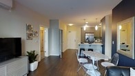 This single-floor residence showcases a thoughtfully designed open-concept layout that seamlessly connects the kitchen, dining, and living areas. The kitchen is equipped with light wood cabinetry, white quartz countertops, a stainless steel undermount sink with a pull-down faucet, and a stainless steel refrigerator with an ice dispenser. Pendant lighting hangs above the kitchen island, which doubles as a casual dining counter with bar seating.

The bold navy blue accent walls create a striking visual contrast against the neutral tones throughout the space, lending it a sophisticated, contemporary character. Dark hardwood flooring runs throughout the main living areas, grounding the open floor plan with warmth and texture.

A dedicated dining area features a marble-top round table with modern wire chairs, positioned near the kitchen for easy flow. The living area is styled with coordinating navy furnishings and benefits from natural light provided by large windows dressed in sheer curtains.

A glimpse into the bedroom reveals a mirrored entryway and a continuing navy color palette, maintaining design cohesion across the unit. Overall, the space presents a polished, modern aesthetic with intentional use of color, lighting, and materials.