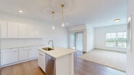 Located at 5155 Raglan St. in Hilliard, Ohio, this well-designed one-bedroom, one-bathroom residence offers a thoughtfully organized layout across a single floor. The open-concept kitchen serves as the centerpiece of the living area, featuring crisp white cabinetry, a tiled backsplash, stainless steel appliances — including a refrigerator, range, and microwave — and a central island with an integrated sink and light quartz countertop. Two gold pendant lights hang above the island, adding a warm, contemporary accent. Light wood-style flooring flows throughout the main living areas, enhancing the bright, clean aesthetic. The bedroom is a comfortable and well-lit retreat with plush carpeting, a large window offering outdoor views, and direct access to a generously sized walk-in closet equipped with wire shelving on multiple walls, providing ample storage. The full bathroom is finished in a clean, all-white palette, featuring a tub-shower combination, a modern vanity, and wood-style flooring that complements the rest of the home. A private balcony rounds out the unit, offering an outdoor extension of the living space. The overall design is modern and neutral, making it a functional and move-in ready residence.