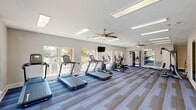This fitness center in Slidell, Louisiana, is a spacious, single-floor facility designed to accommodate a comprehensive workout experience. The main exercise area is generously proportioned, featuring striped, multi-tone carpet flooring in blue and gray hues that runs throughout the space. Large windows line the perimeter walls, flooding the interior with natural light and offering views of the surrounding outdoor areas, including what appears to be a pool terrace. Ceiling fans are positioned throughout to promote air circulation and comfort during workouts.

The cardio zone is well-stocked with multiple treadmills and elliptical machines arranged in a row along the windowed wall, complemented by wall-mounted flat-screen televisions for entertainment. A multi-station cable machine and a weight bench provide strength training options, while a dedicated dumbbell rack offers a range of free weights. Large framed mirrors are installed along one wall, enhancing the sense of space and allowing for form monitoring during exercise.

A separate spin room is included, equipped with stationary cycling bikes and its own mounted televisions, providing a focused environment for cycling workouts. Recessed panel lighting throughout ensures bright, even illumination. The overall ambiance is clean, modern, and functional.