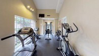 This fitness center offers a comprehensive and welcoming environment for exercise and wellness. The space is defined by its notably high ceilings, which lend an open, airy quality throughout, and large multi-pane windows that flood the interior with natural light and provide views of the surrounding outdoor greenery. The flooring is finished in durable dark rubber matting, practical for a high-activity environment.

The main workout area is thoughtfully organized to accommodate a variety of training styles. Cardio equipment includes treadmills, an upright stationary bike, and elliptical trainers, all positioned to take advantage of the natural light from the large windows. A multi-station cable machine anchors the strength training section, complemented by a full rack of dumbbells in graduated weights and a flat workout bench, catering to both beginners and experienced users.

Track lighting along the upper walls provides focused illumination, enhancing the warm, neutral tones of the painted walls. Workout reference charts are posted for guidance on training techniques and heart rate zones. A glass-paned door connects to an adjacent area, suggesting additional amenity spaces nearby. A hand sanitizer dispenser near the entrance reflects attention to cleanliness and user convenience. Overall, the fitness center presents a functional, well-maintained space designed to support a well-rounded exercise routine.