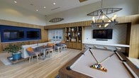 This single-floor amenity lounge delivers a well-rounded recreational experience through a thoughtfully designed, open-plan layout. The space is anchored by a full-sized pool table at the forefront, setting an immediately social and engaging tone. Warm hardwood-style flooring runs throughout, complementing the rich wood wall paneling that lines much of the interior and contributes to a cohesive coastal aesthetic.

A built-in shelving unit along the back wall provides both decorative display space and functional storage, flanked by a wall-mounted flat-screen television. A second large TV is positioned on the opposite wall, ensuring clear sightlines from multiple seating areas. The lounge seating area features a generous sectional sofa accented with colorful orange and teal throw pillows, paired with round café-style tables and mixed dining chairs that encourage casual gathering.

Overhead, a statement chandelier with exposed Edison bulbs and a nautical rope design serves as a striking focal point, reinforcing the coastal theme. Decorative touches — including a canoe-shaped wall piece and herringbone-patterned accent walls — add visual interest throughout. The elevated ceiling height enhances the overall sense of openness, making this a versatile and inviting communal space.