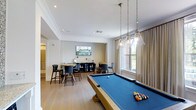 This single-floor amenity lounge is thoughtfully designed to offer a welcoming and versatile social space. At the heart of the room sits a full-size billiards table with a rich blue felt surface, suspended above by elegant pendant lights with glass shades that cast warm, focused illumination. The light hardwood flooring runs throughout, complementing the bright, neutral color palette of the walls and crown-molded ceilings. Recessed lighting adds to the layered lighting scheme, enhancing the room's polished yet relaxed atmosphere. A dedicated seating area is arranged toward the rear of the space, featuring bar-height tables and chairs alongside a wall-mounted flat-screen TV, creating an ideal setting for casual conversation or watching a game. A dark wood credenza anchors the seating nook, adding a touch of sophistication. Large windows dressed with floor-length linen drapes allow natural light to filter in, offering views of the surrounding greenery while maintaining a sense of privacy. The open layout ensures ease of movement throughout, making the space equally suitable for intimate gatherings or larger social events. Overall, the lounge strikes a balance between functional design and refined comfort.