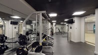 This fitness facility is designed to support a full range of strength and conditioning workouts within a thoughtfully organized, single-level layout. The space is divided into distinct workout zones, each outfitted with professional-grade equipment. A dedicated free weights area features Paramount cable machines, squat racks, barbells, weight plates, and multiple adjustable and flat benches, accommodating a wide variety of resistance training exercises. A separate section houses additional cable and functional training stations, including a resistance band tower and multi-functional cable systems. A dumbbell rack with a selection of weights and adjustable benches rounds out the strength equipment offerings.

The interior design features a striking black ceiling that contrasts with light grey walls, creating a focused and modern training environment. Floor-to-ceiling mirrors line the walls throughout, visually expanding the space while allowing users to monitor their form. Durable speckled rubber flooring provides traction and impact absorption across the entire facility. Recessed panel lighting ensures consistent, even illumination. A wall-mounted flat-screen television is positioned for visibility across the main training area. A glass partition with a secondary door connects the gym to an adjacent space, providing a seamless transition between areas.