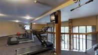 This fitness center offers a practical and well-appointed space designed for cardio-focused workouts. The room features a lineup of cardio equipment, including treadmills and elliptical machines, arranged along a railed balcony-style perimeter that allows natural light to flow in through large windows, providing views of the surrounding greenery outside. A wall-mounted flat-screen television is positioned on a structural wood post, keeping exercisers entertained during their workouts. The interior design blends warm wood beam accents and exposed brick columns with a neutral color palette, contributing to a welcoming yet functional atmosphere. The textured ceiling and flush-mounted lighting complement the overall aesthetic, while an exit sign indicates clear safety provisions throughout the space. The open layout ensures ample room to move between equipment, and the railing detail along the open edge adds both a safety feature and a distinctive architectural element. This fitness center is well-suited for residents or guests seeking a convenient, comfortable space for their daily exercise routines.