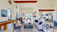 Located at 101 Barnes Ln in Broussard, Louisiana, this single-story community clubhouse offers a generously sized, open-plan gathering space designed with a clean, contemporary aesthetic. The interior is defined by dramatically soaring ceilings accented by rich, dark wood beams that contrast beautifully against crisp white walls, creating an airy and inviting atmosphere throughout.

The main lounge area features large-format tile flooring and is furnished with comfortable seating arrangements centered around a rustic wood coffee table, ideal for socializing or relaxing. A full-sized billiards table anchors the game area, which is bathed in natural light streaming through floor-to-ceiling black-framed glass walls and clerestory windows. These expansive glazed panels provide direct sightlines and access to the outdoor pool area beyond.

The entry vestibule makes a strong first impression with its two-story curtain wall of black-framed glass, flooding the space with abundant natural light and offering open views of the surrounding landscaping.

The kitchen is well-appointed with slate-blue shaker cabinetry, white quartz countertops, stainless steel appliances, and a large central island with bar seating, all illuminated by stylish globe pendant lights. A dining area with round tables and upholstered chairs completes the space, making this clubhouse a fully functional and welcoming community hub.