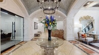 Located at 3500 Fairmount St in Dallas, Texas, this sophisticated lobby space exudes classical elegance through its thoughtfully curated architectural details. The grand entrance is anchored by a stunning circular glass-topped pedestal table, adorned with a dramatic floral arrangement beneath an ornate wrought-iron chandelier. The vaulted ceiling above features an intricate mosaic tile treatment that spans the arched entryway, creating a richly textured focal point.

Warm-toned marble tile flooring with decorative inlay patterns flows throughout, complementing the rich, multi-veined marble reception desk that commands attention near the center of the space. Crisp white wainscoting and paneled millwork line the walls, accented by diamond-pattern details that reinforce the refined aesthetic. Flanking the arched hallway corridor are two stately wrought-iron carriage-style wall lanterns that guide visitors deeper into the space.

The hallway leads past rich wood-toned doors to additional rooms, framed by a graceful barrel-vaulted ceiling with mosaic accents. An elegant staircase with ornate iron balusters connects to the upper level. A comfortable seating area with French-style upholstered chairs and warm accent pillows adds a welcoming touch, completing an environment that balances grandeur with approachable sophistication.
