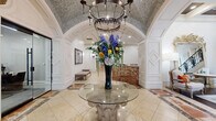 Located at 3500 Fairmount St in Dallas, Texas, this sophisticated lobby space exudes classical elegance through its thoughtfully curated architectural details. The grand entrance is anchored by a stunning circular glass-topped pedestal table, adorned with a dramatic floral arrangement beneath an ornate wrought-iron chandelier. The vaulted ceiling above features an intricate mosaic tile treatment that spans the arched entryway, creating a richly textured focal point.

Warm-toned marble tile flooring with decorative inlay patterns flows throughout, complementing the rich, multi-veined marble reception desk that commands attention near the center of the space. Crisp white wainscoting and paneled millwork line the walls, accented by diamond-pattern details that reinforce the refined aesthetic. Flanking the arched hallway corridor are two stately wrought-iron carriage-style wall lanterns that guide visitors deeper into the space.

The hallway leads past rich wood-toned doors to additional rooms, framed by a graceful barrel-vaulted ceiling with mosaic accents. An elegant staircase with ornate iron balusters connects to the upper level. A comfortable seating area with French-style upholstered chairs and warm accent pillows adds a welcoming touch, completing an environment that balances grandeur with approachable sophistication.