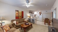 This one-bedroom, one-bathroom unit in Fayetteville, Arkansas offers a thoughtfully arranged single-level floor plan that maximizes functionality. The combined living and kitchen area forms the heart of the space, where a ceiling fan overhead and natural light from a large window create a bright, comfortable atmosphere. The living area features neutral carpet and ample room for seating arrangements, while the adjoining kitchen is equipped with white cabinetry extending to the ceiling, a full appliance suite including a refrigerator, electric range, built-in microwave, and dishwasher, plus a peninsula counter with bar seating that serves as a natural divider between the two spaces. Tile flooring runs throughout the kitchen for easy maintenance. The bedroom offers a generous footprint with carpet underfoot, a ceiling fan with light fixture, a large window that brings in natural light, and access to a dedicated closet. The bathroom presents a clean, efficient design with a white vanity, mirror, tile flooring, and overhead lighting. A separate closet near the kitchen provides additional storage. The overall aesthetic is neutral and functional, with consistent finishes throughout.