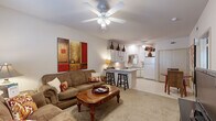 This one-bedroom, one-bathroom unit in Fayetteville, Arkansas offers a thoughtfully arranged single-level floor plan that maximizes functionality. The combined living and kitchen area forms the heart of the space, where a ceiling fan overhead and natural light from a large window create a bright, comfortable atmosphere. The living area features neutral carpet and ample room for seating arrangements, while the adjoining kitchen is equipped with white cabinetry extending to the ceiling, a full appliance suite including a refrigerator, electric range, built-in microwave, and dishwasher, plus a peninsula counter with bar seating that serves as a natural divider between the two spaces. Tile flooring runs throughout the kitchen for easy maintenance. The bedroom offers a generous footprint with carpet underfoot, a ceiling fan with light fixture, a large window that brings in natural light, and access to a dedicated closet. The bathroom presents a clean, efficient design with a white vanity, mirror, tile flooring, and overhead lighting. A separate closet near the kitchen provides additional storage. The overall aesthetic is neutral and functional, with consistent finishes throughout.